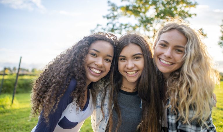 A small group of three female friends huddle in closely together as they pose for a portrait.  They are each dressed casually and are smiling on the warm summers evening.