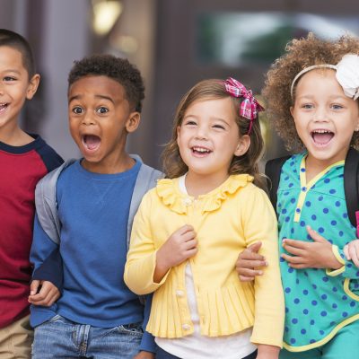 A multi-ethnic group of children standing in a row in a school hallway, excited and laughing, watching something.  They are in kindergarten or preschool, carrying bookbags.  They are 4 to 6 years old.