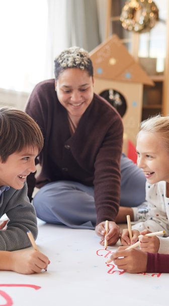 Full length portrait of smiling female teacher sitting on floor with multi-ethnic group of kids drawing pictures while enjoying art class, copy space